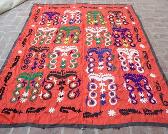 Könnte beinhalten: Ein großes, rechteckiges, rotes Textil mit einem sich wiederholenden Muster aus stilisierten floralen und geometrischen Formen in Weiß, Grün, Blau, Lila und Schwarz. Das Textil hat einen dunkelgrauen Rand und ist wahrscheinlich ein dekorativer Teppich oder Wandbehang.