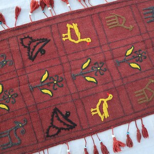 May include: A red woven rug with a geometric pattern of birds, plants, and animals. The rug has fringe on all four sides.