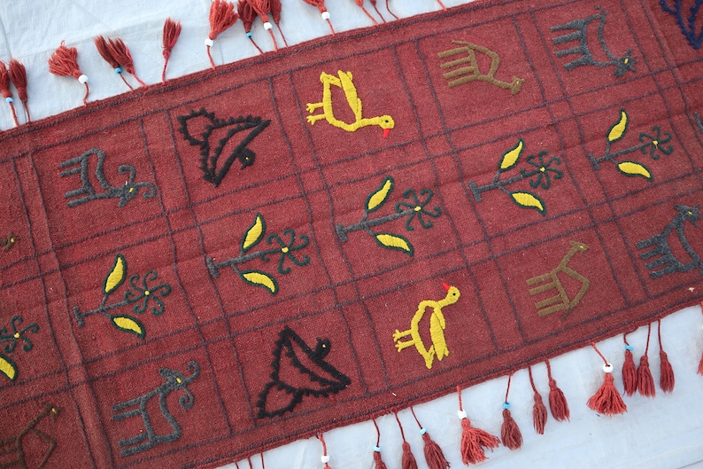 May include: A red woven rug with a geometric pattern of birds, plants, and animals. The rug has fringe on all four sides.
