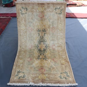 May include: A rectangular beige rug with a floral pattern in shades of pink, green, and yellow. The rug has a detailed design with a central medallion and a border. The edges are finished with a fringe.