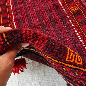 May include: A hand-knotted rug featuring a deep red base with intricate black and orange geometric patterns. The rug has a thick pile texture and is finished with decorative tassels along the edges, which are adorned with small blue and white beads.