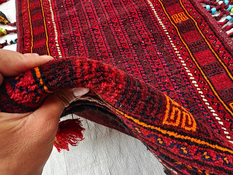 May include: A hand-knotted rug featuring a deep red base with intricate black and orange geometric patterns. The rug has a thick pile texture and is finished with decorative tassels along the edges, which are adorned with small blue and white beads.