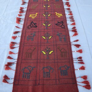 May include: A red wool rug with a geometric pattern of animals and plants. The rug has fringe on all four sides and is decorated with tassels.