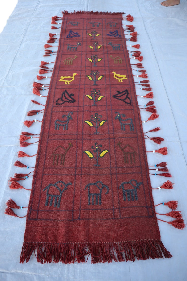 May include: A red wool rug with a geometric pattern of animals and plants. The rug has fringe on all four sides and is decorated with tassels.