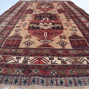 Pu&ograve; includere: Un tappeto tradizionale con sfondo beige e intricati motivi in rosso, nero e verde. Il tappeto presenta un disegno centrale con un vaso e motivi floreali.