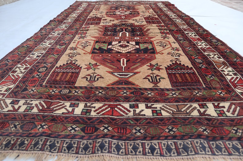 Pu&ograve; includere: Un tappeto tradizionale con sfondo beige e intricati motivi in rosso, nero e verde. Il tappeto presenta un disegno centrale con un vaso e motivi floreali.