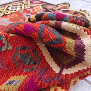 Op de afbeelding: Een geweven kleed met een levendig geometrisch patroon in tinten rood, oranje, geel, donkerbruin, blauw en magenta. Het textiel vertoont een gevouwen hoek, die een dik, getextureerd weefsel met ingewikkelde ruit- en zigzagmotieven onthult.