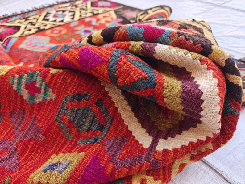 Op de afbeelding: Een geweven kleed met een levendig geometrisch patroon in tinten rood, oranje, geel, donkerbruin, blauw en magenta. Het textiel vertoont een gevouwen hoek, die een dik, getextureerd weefsel met ingewikkelde ruit- en zigzagmotieven onthult.