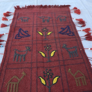 May include: A red woven rug with a geometric pattern of animals and flowers. The rug has a fringe border with tassels.