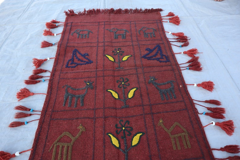 May include: A red woven rug with a geometric pattern of animals and flowers. The rug has a fringe border with tassels.