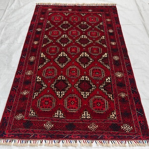 May include: A rectangular area rug featuring a deep red background with a repeating pattern of dark navy and cream geometric diamond and octagonal motifs. The rug is framed by a wide border with intricate linear designs and short white fringe.
