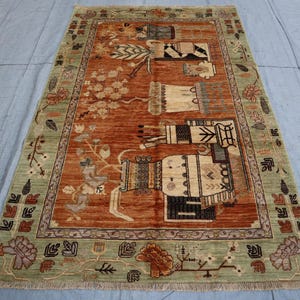 May include: A rectangular rug with a central rust-coloured field featuring stylised figures and floral motifs. The border is a light green with repeating patterns. The rug's design includes beige, black, and brown elements, creating a detailed and decorative piece.