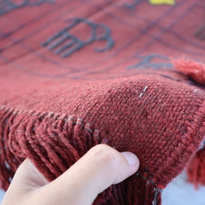 May include: Close-up of a red woven rug with fringe. The rug has a geometric pattern with black lines and a few yellow accents.