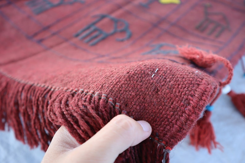 May include: Close-up of a red woven rug with fringe. The rug has a geometric pattern with black lines and a few yellow accents.