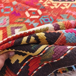 Op de afbeelding: Een geweven kleed met een levendig geometrisch patroon in tinten rood, oranje, geel, blauw, paars en zwart. Het textiel vertoont een dicht, ingewikkeld ontwerp met herhalende hoekige motieven en een dikke, getextureerde weving in de vouw.