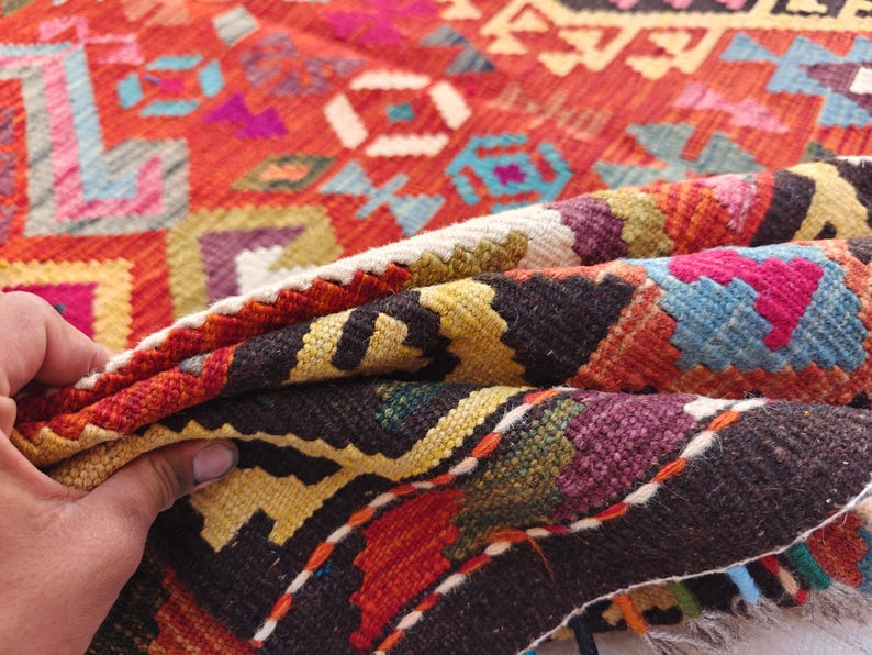 Op de afbeelding: Een geweven kleed met een levendig geometrisch patroon in tinten rood, oranje, geel, blauw, paars en zwart. Het textiel vertoont een dicht, ingewikkeld ontwerp met herhalende hoekige motieven en een dikke, getextureerde weving in de vouw.