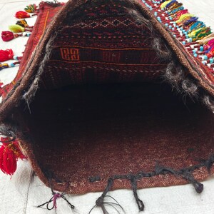 May include: A woven textile bag featuring a dark brown and black geometric pattern on the interior flap. The exterior edges are adorned with numerous multi-colored tassels in shades of red, yellow, blue, and pink, accented with small beads.