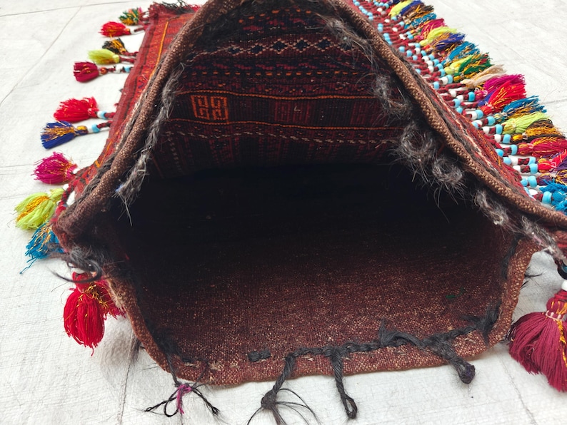 May include: A woven textile bag featuring a dark brown and black geometric pattern on the interior flap. The exterior edges are adorned with numerous multi-colored tassels in shades of red, yellow, blue, and pink, accented with small beads.
