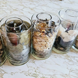 May include: Six clear glass jars filled with various seashells and other beach finds. The jars are arranged in a row on a white marble surface.