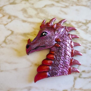 May include: A pink and red dragon head sculpture with a textured surface and a spiky mane. The dragon has a single black eye and a detailed snout.