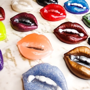 May include: A collection of 15 colorful, glossy, and glittery lips with teeth, made of resin. The lips are in various colors, including red, pink, purple, green, blue, black, and yellow.