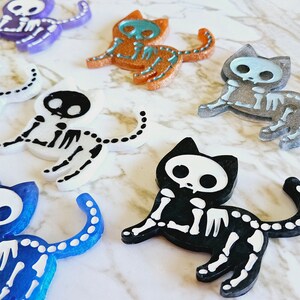 May include: A collection of cat-shaped resin figurines with skeleton designs. The figurines are in various colors, including white, black, blue, orange, gray, and purple. Each cat has a detailed skeleton pattern and a glossy finish, perfect for Halloween decor.