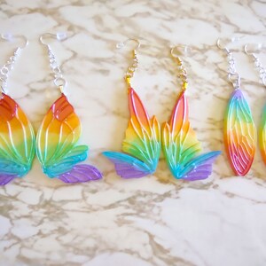 May include: A set of four pairs of rainbow-colored butterfly earrings. The earrings are made of clear plastic and have a holographic finish. The butterfly wings are shaped like a teardrop and have a rainbow gradient.