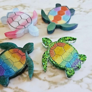 May include: Four resin turtle ornaments with a hexagonal shell design. Each turtle has a different color scheme: pink and white, orange and yellow, green and blue, and rainbow.