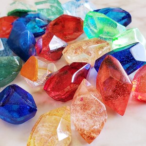 May include: A collection of colorful, faceted, glass gems in various shades of red, blue, green, orange, and yellow. The gems are arranged on a white marble surface.