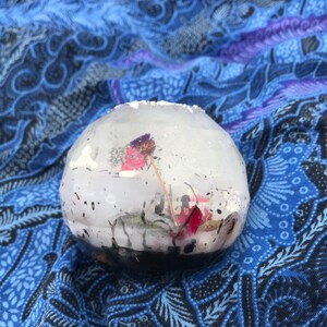 Könnte beinhalten: Eine klare Glaskugel mit einer getrockneten Blume und einem Stiel darin. Die Kugel liegt auf einem blauen und violetten gemusterten Stoff.