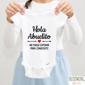 May include: White baby onesie with black text that reads "Hola Abuelito" with a red heart and arrows. The text below reads "No puedo esperar para conocerte".