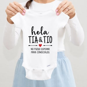 May include: White baby onesie with black text that reads "hola tia & tio" with a red heart and the text "no puedo esperar para conocerlos" below.