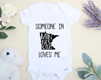 May include: White baby onesie with black text that reads "Someone in Minnesota loves me". The text is arranged in the shape of the state of Minnesota.