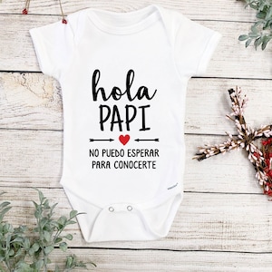 May include: White baby onesie with black text that reads "hola papi" with a red heart and the text "no puedo esperar para conocerte".
