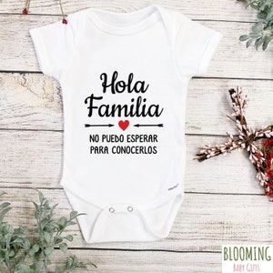 May include: White baby onesie with black text that reads "Hola Familia" with a red heart and arrows pointing to the text. The text below the arrows reads "No puedo esperar para conocerlos".