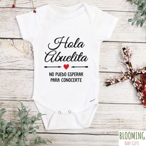 May include: White baby onesie with black text that reads "Hola Abuelita" with a red heart and arrows. The text below reads "No puedo esperar para conocerte".