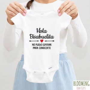 May include: White baby onesie with black text that reads "Hola Bisabuelita" with a red heart and arrows pointing to the text. The text below the arrows reads "No puedo esperar para conocerte".