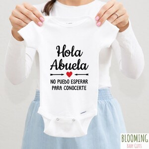 May include: White baby onesie with black text that reads "Hola Abuela" with a red heart and arrows pointing to the text. The text below reads "No puedo esperar para conocerte".