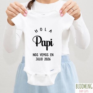 May include: White baby onesie with black text that reads "Hola Papi Nos Vemos en Julio 2026".