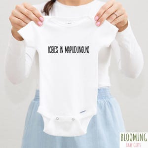 May include: White baby onesie with the text "(CRIES IN MAPUDUNGUN)" in black. The onesie is held up by a person. The Blooming Baby Gifts logo is in the bottom right corner.