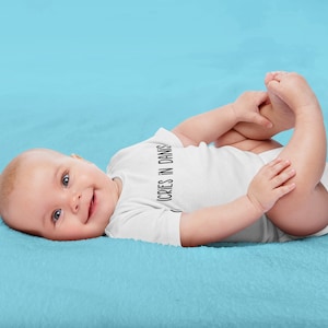 May include: A baby wearing a white onesie with the text "CRIES IN DANISH" on it, lying on a light blue blanket. The baby is smiling and looking at the camera.