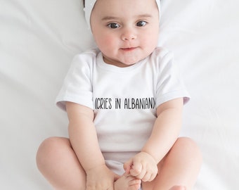 May include: A white onesie with the text "(CRIES IN ALBANIAN)" on the front. A baby wearing the onesie is lying on a white sheet. The baby is wearing a white headband with a heart-shaped bow.