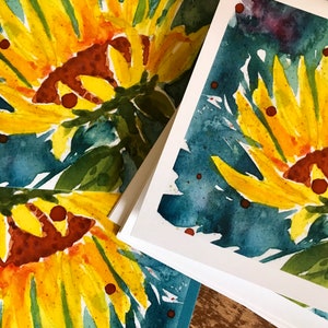 May include: A stack of greeting cards with a watercolor painting of sunflowers on a blue background. The sunflowers are yellow and orange with brown centers.