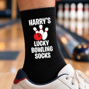 May include: A black crew sock featuring white text that reads HARRY'S LUCKY BOWLING SOCKS. Above the text is a graphic of a red bowling ball and three white bowling pins with red stripes. The sock is worn inside a white athletic shoe.