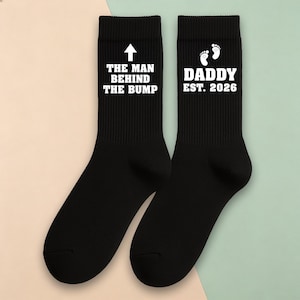 Pode incluir: Meias pretas com texto e gráficos brancos. Uma meia diz "THE MAN BEHIND THE BUMP" com uma seta. A outra diz "DADDY EST. 2026" com pegadas. Um presente divertido para futuros pais.