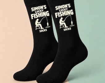May include: A pair of black crew socks featuring a white graphic of a person sitting on a stool fishing with a rod and line. The text printed in white reads SIMON'S LUCKY FISHING SOCKS, with the graphic positioned between the words LUCKY and SOCKS.