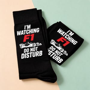 May include: A pair of black crew socks featuring white text that reads I'M WATCHING F1 DO NOT DISTURB with the F1 in red. A white graphic of a Formula 1 race car is positioned between the lines of text on the front of each sock.