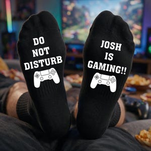 May include: A pair of black socks featuring white text and a white game controller graphic. The left sock reads DO NOT DISTURB and the right sock reads JOSH IS GAMING!!, both printed in a bold, sans-serif font above the controller icon.
