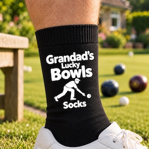 May include: A black crew sock featuring white text that reads Grandad's Lucky Bowls Socks, accompanied by a white graphic illustration of a person in a crouching position preparing to roll a lawn bowl ball on a green grass surface.