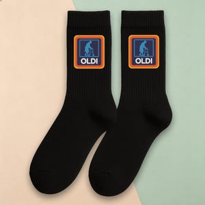 May include: A pair of black crew socks featuring a square graphic on the upper portion. The graphic displays a blue silhouette of a person using a walker inside a frame with orange and yellow borders, with the white text OLDI printed below.
